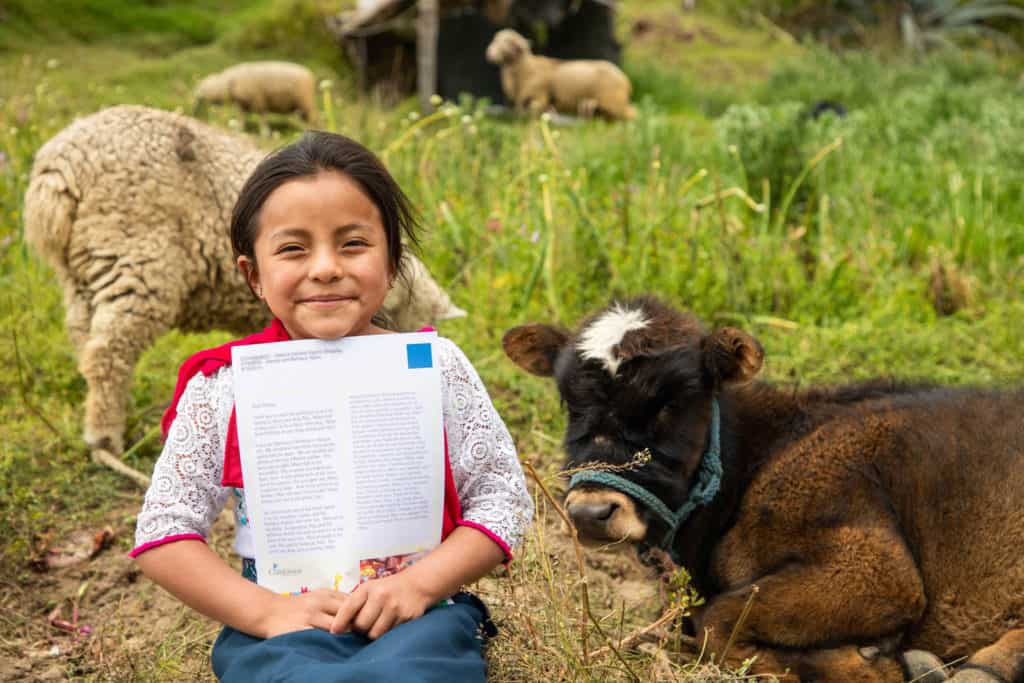 A girl sits in a grassy field with sheep and a cow behind her, holding a letter from her sponsor and smiling
