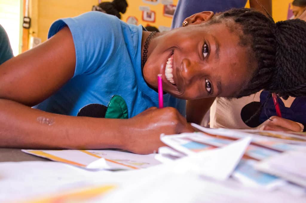 A girl wearing a blue shirt, writing letters to her sponsor. Letters from her sponsor surround her as she sits at the table.  She is leaning in close and smiling.