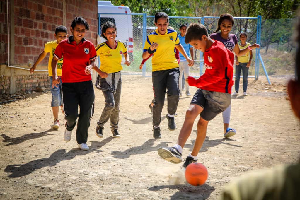 A group of boys and girls are playing soccer on a dirt field at the project. A boy in a red shirt is kicking the ball.