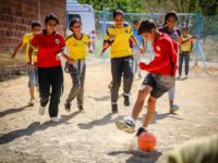 A group of boys and girls are playing soccer on a dirt field at the project. A boy in a red shirt is kicking the ball.