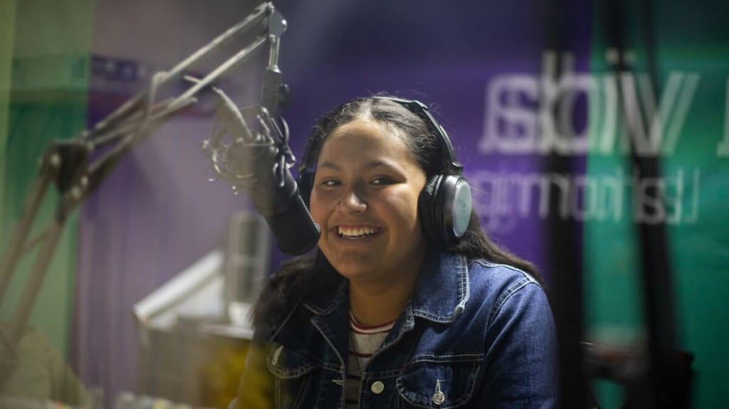 a teen girl wears headphones and sits behind the microphone of a radio station. She is wearing a denim jacket and smiling at the camera