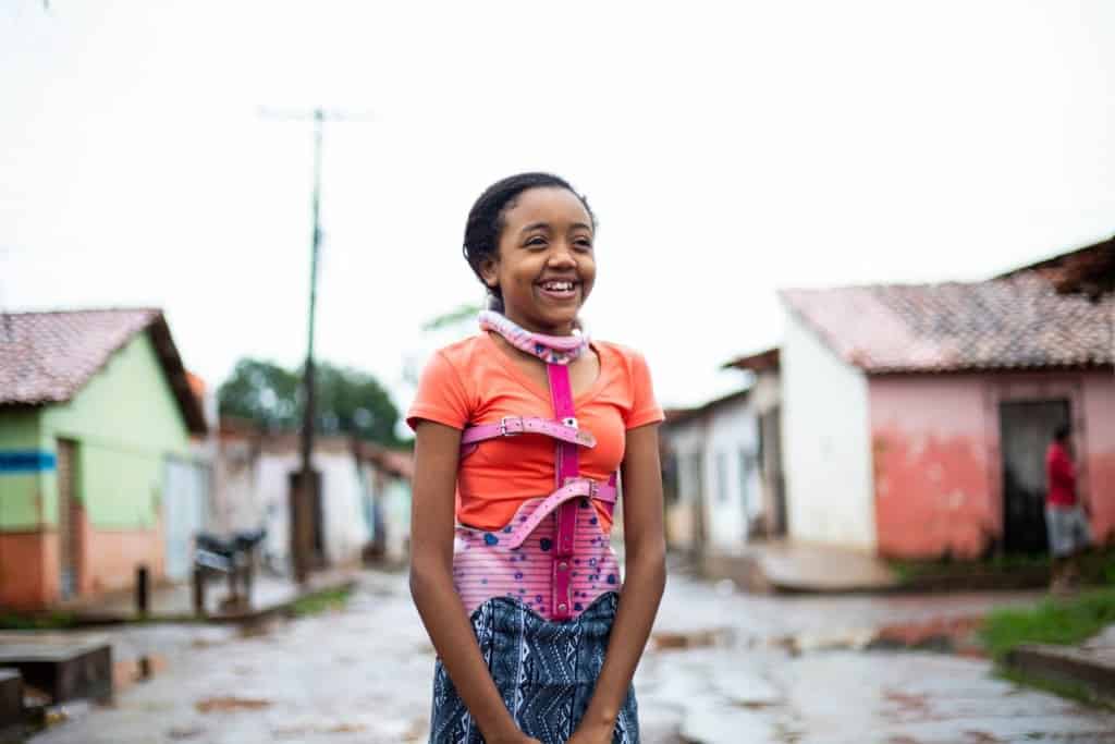 A teen girl stands outside smiling. She is wearing colorful clothing and a back brace over her clothing.