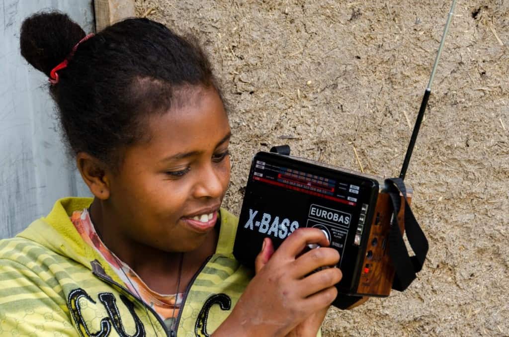 A teen girl holds a radio and tunes a dial. She is wearing a yellow shirt and sitting in front of a mud home.