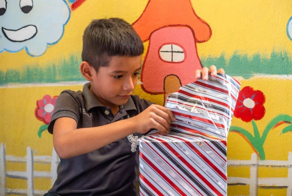 A boy wearing a gray shirt sits in front of a colorfully painted wall, opening a square Christmas gift wrapped in shiny paper.