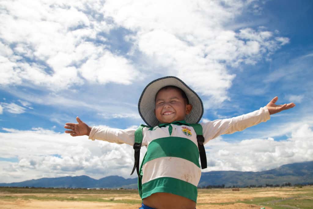 José Luis is wearing a green and white shirt, a hat, and a backpack. He is standing outside his with his arms outstretched. 