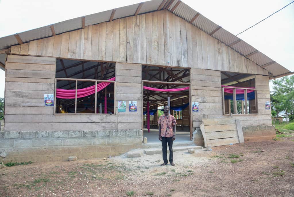 Pastor Joseph is standing in front of the new building. He is wearing dark pants and a pink patterned shirt.