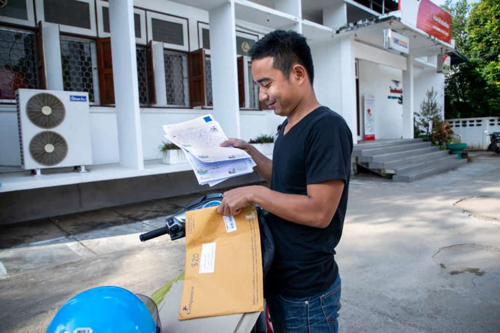 A man wearing a black shirt places child letters into a manila envelope. He is standing outside near a motorbike.