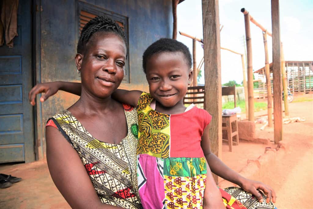 Sabina and her mother are both looking at the camera and smiling, wearing colorful shirts. The background is a blue building. Sabina has her arm around her mother.