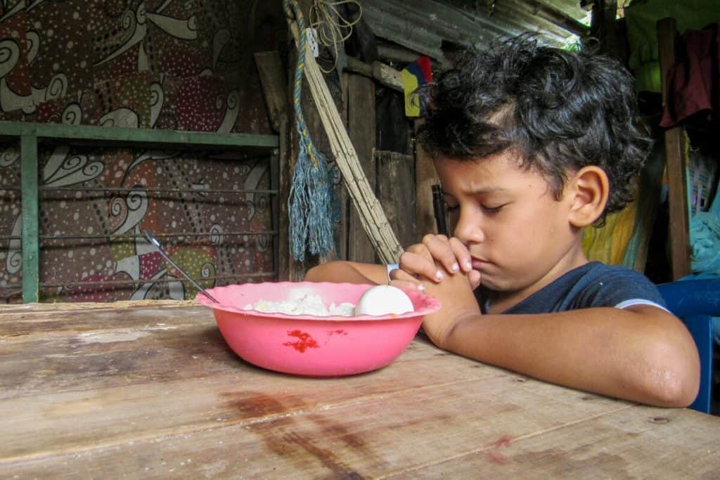 A young boy in Colombia closes his eyes and folds his hands in prayer before a meal. There is a pink bowl with food in it on a table in front of him.