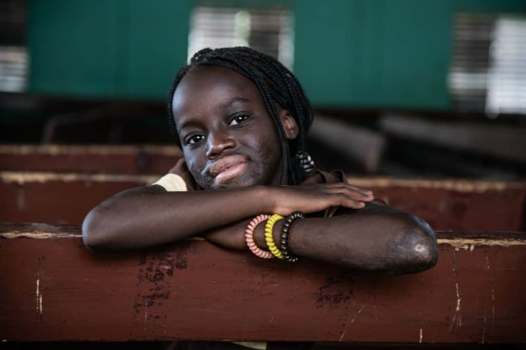 A girl rests her arms and head on a wooden bench and smiles. She is wearing three bracelets.