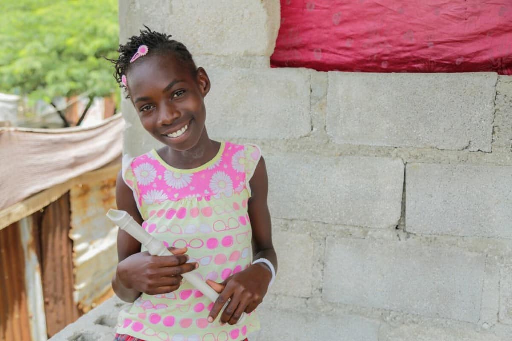 a 12-year-old girl in pink and white shirt smiles. She is standing in front of a concrete wall and holding a recorder instrument