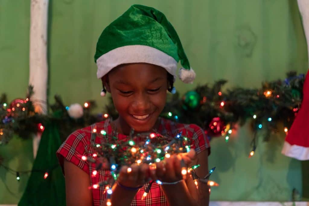 An adolescent girl wearing a green Santa hat and plaid red dress holds a bundle of colorful Christmas lights