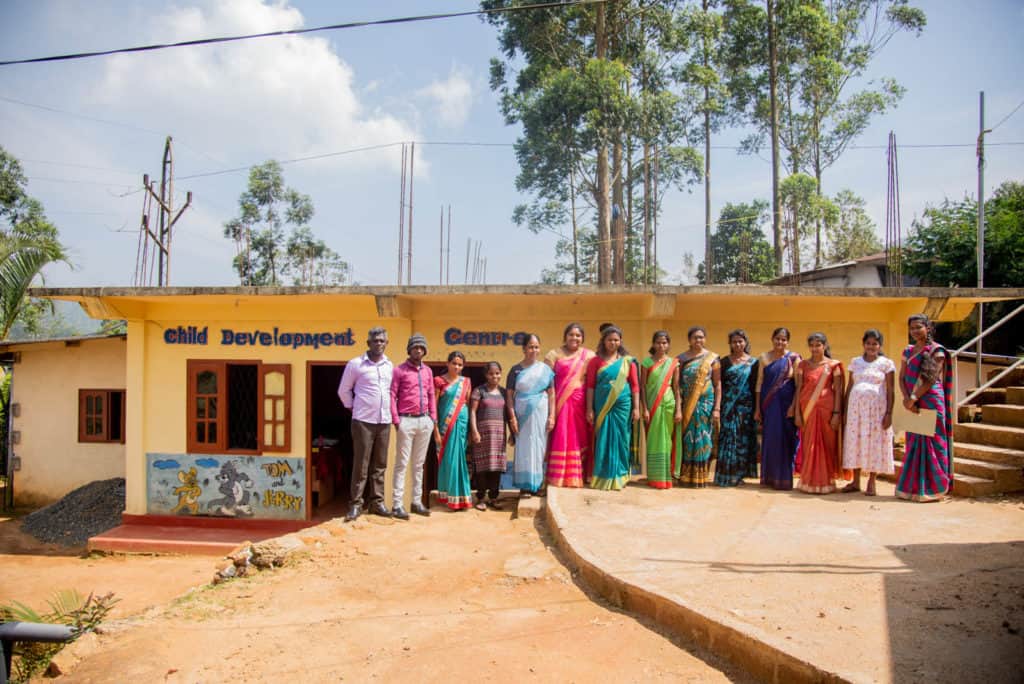 They are posing for a picture in front of the Child Development Center building. Pastor Babu, in a lavender shirt, is on the far left.