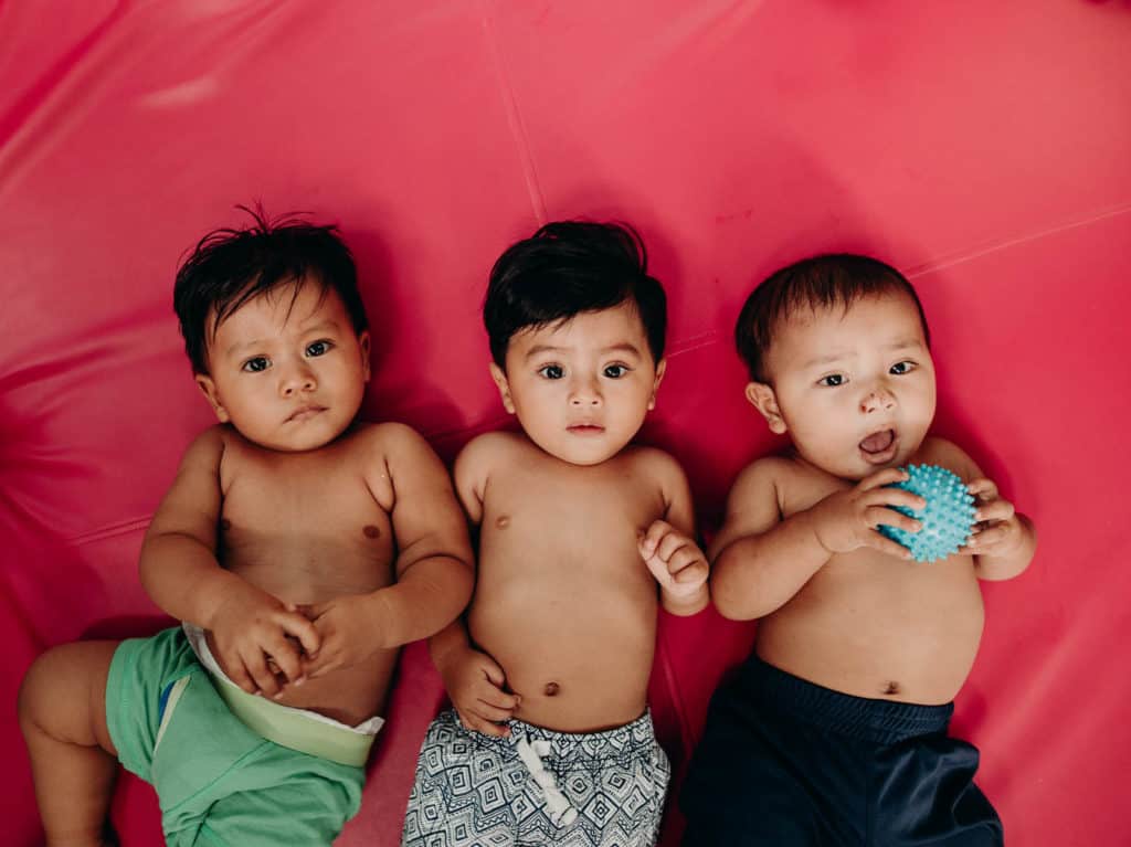 From left to right, Delvin, in green shorts, Axel and Axel lie on their backs on a red mat on the floor in the playroom at the child development center.