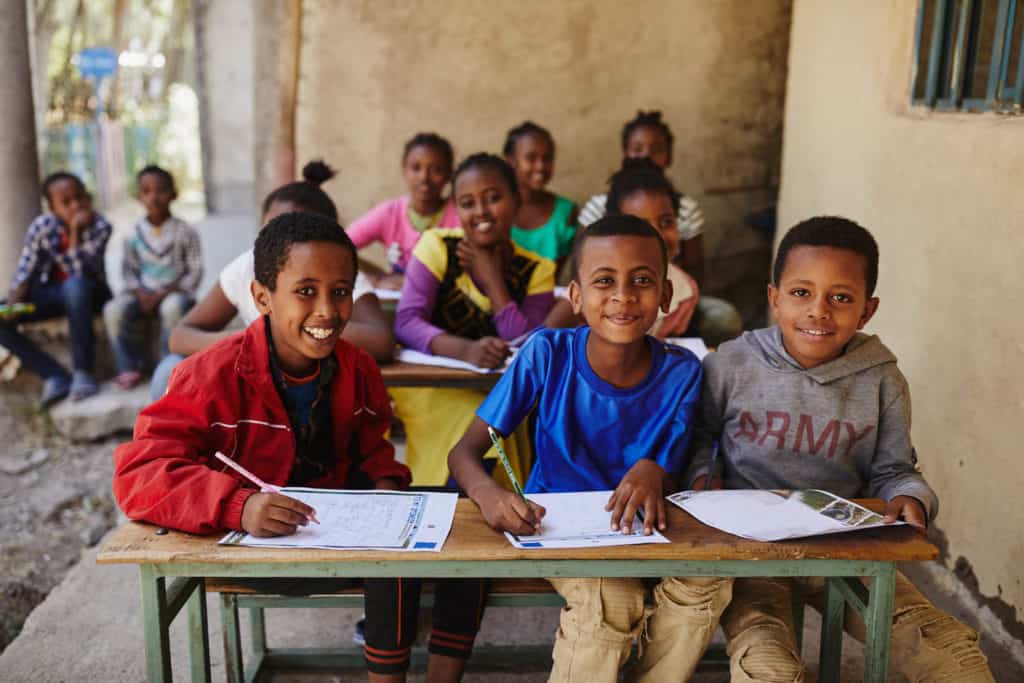 Three boys sit at a desk outside. They  have letters on the desk in front of them, preparing to write. Two rows of children sit at desks behind them. 