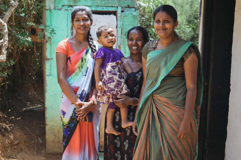 Pavishana is wearing a dress with a purple bodice and a yellow and purple floral print skirt. Her mother, Malathy, wearing a black dress with red and white designs on it, is holding her. They are standing with Pramila, her Project Implementer, and Angel, the Project Manager. All of the women are outside Malathy's home.