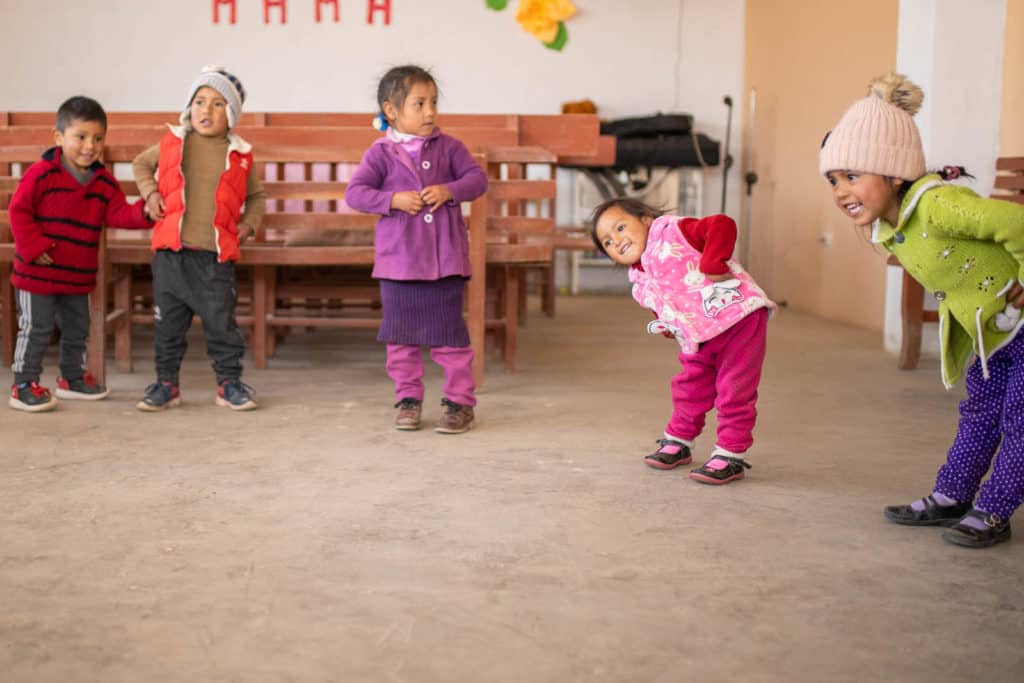 Genesis is wearing red pants with black polka dots, a red shirt and a pink vest. She is in a large classroom playing with her friends. They are dancing.