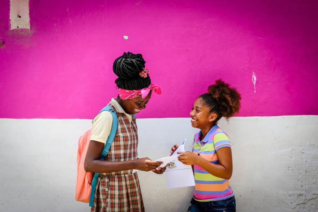A girl wearing a bandana and a girl in a striped shirt are standing in front of a pink and white wall, looking at each other, smiling and laughing. They are holding sponsor letters.