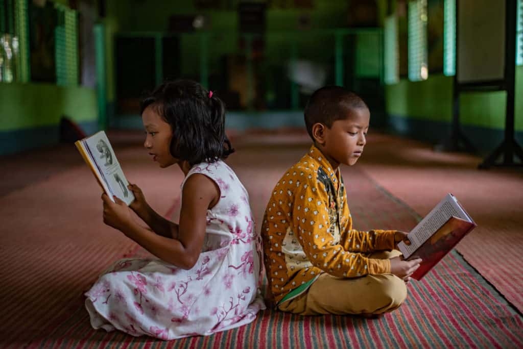 Snigdha, in a white and pink dress, and Prosanto, in a yellow shirt and pants, are sitting on the rug in the activity room holding and reading books.