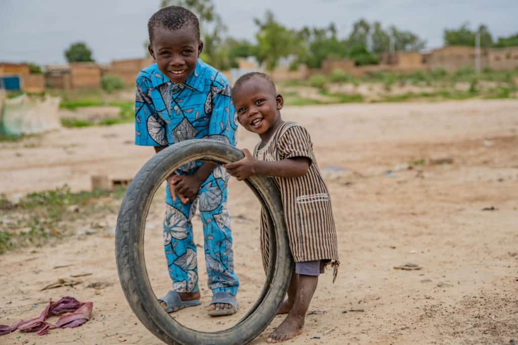 Samsoudine is wearing a blue outfit with black and white designs on it. He is standing outside his home with his younger brother, Adboul, who is holding up an old tire.