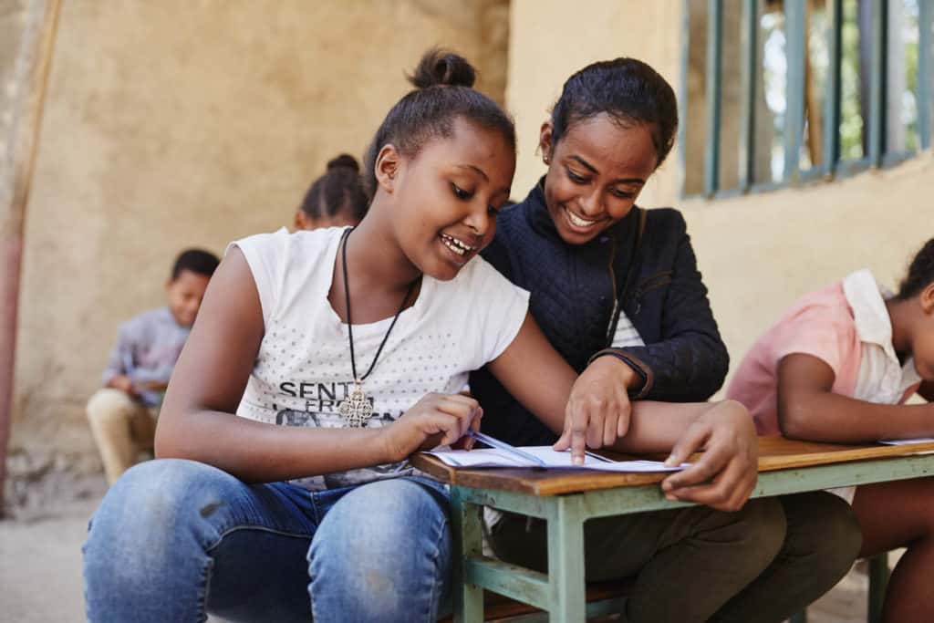 Betey, in a black jacket and white striped shirt, sits at a wood desk working with Yeabsira, in a white shirt and blue jeans, who is writing a letter to her sponsor. There is a tan stucco wall in the background.