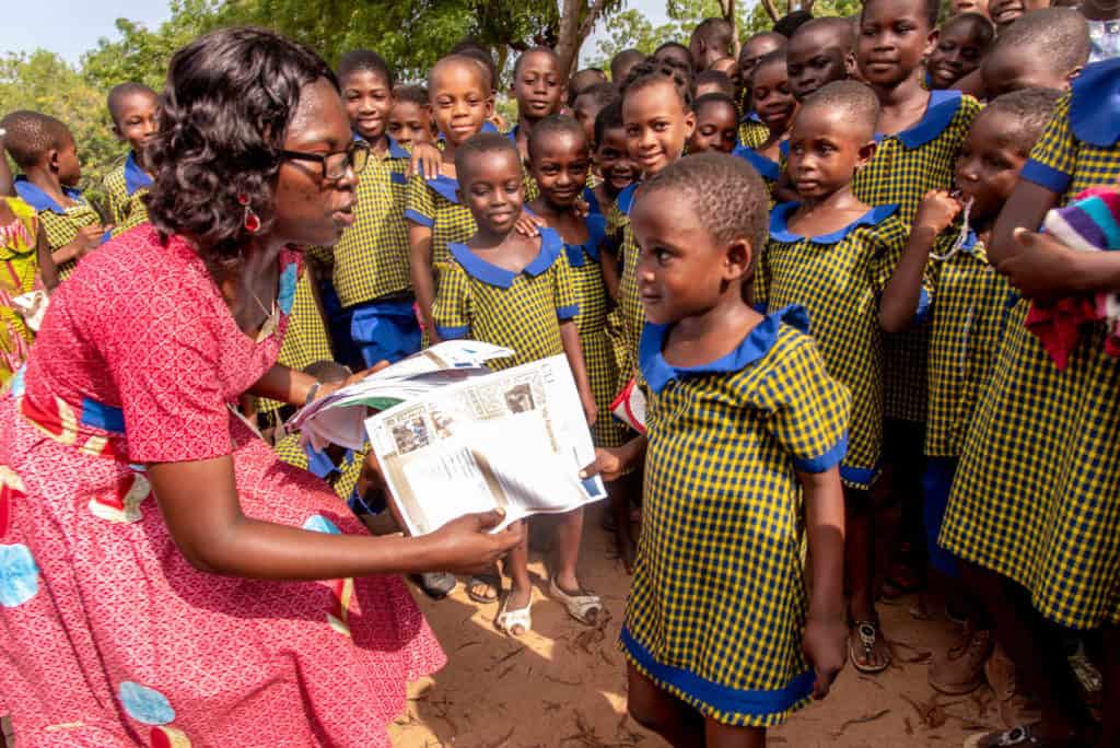 Lorlor, a woman wearing a pink dress, hands out letters from sponsors to children in Ghana. The children are wearing yellow and blue outfits and smiling.