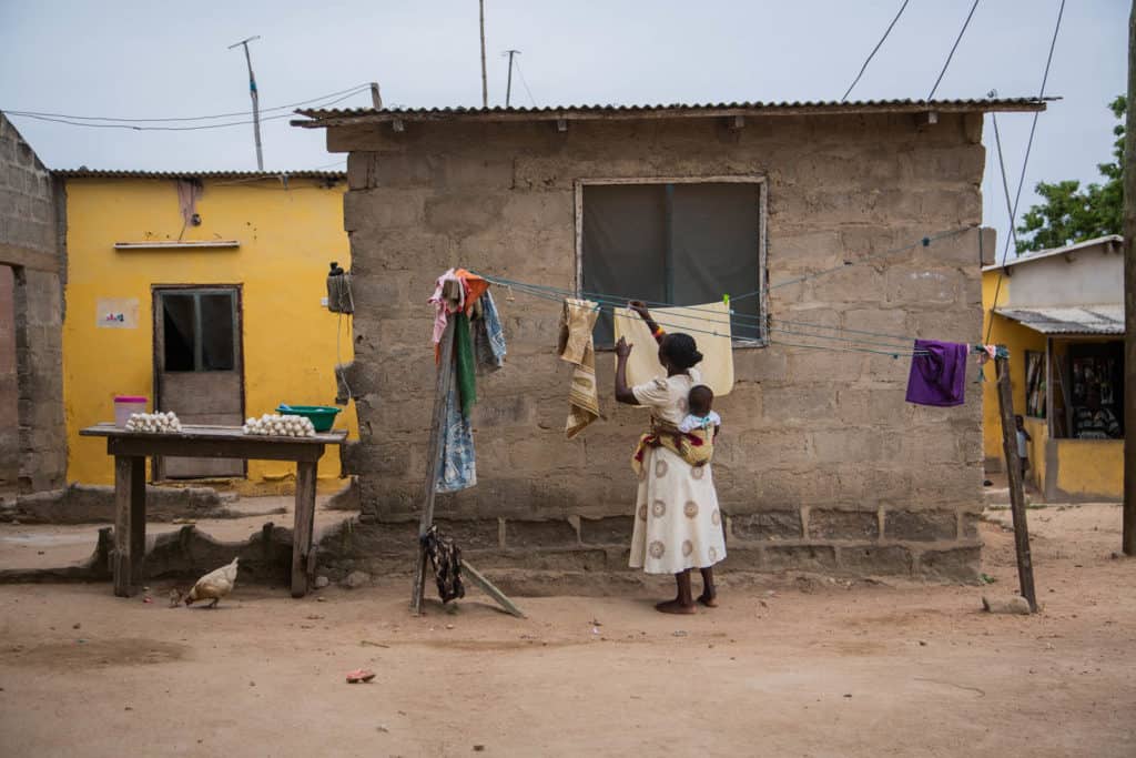 A woman in a white skirt stands outside hanging laundry in front of a brick, cinder block and painted yellow building. She is carrying a baby on her back.