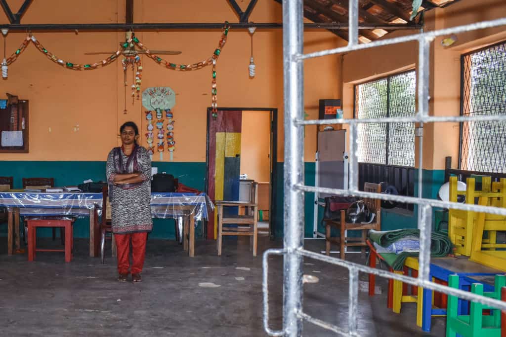 Joanna is wearing red pants with a black and white shirt. She is standing inside the empty Compassion center with her arms crossed in front of her. There is a long table behind her and there are colorful rings of paper hanging above her.