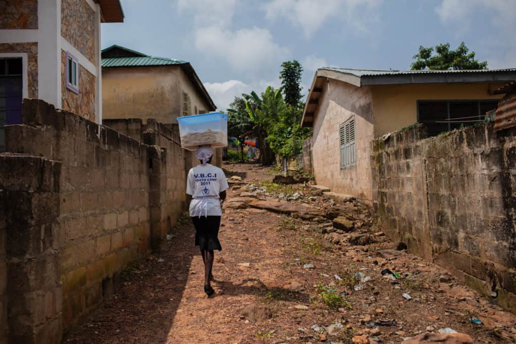 A woman in a white shirt and black skirt is walking up a hill along a dirt path, road between brick walls and is carrying a clear plastic bin with a blue lid on top of her head.