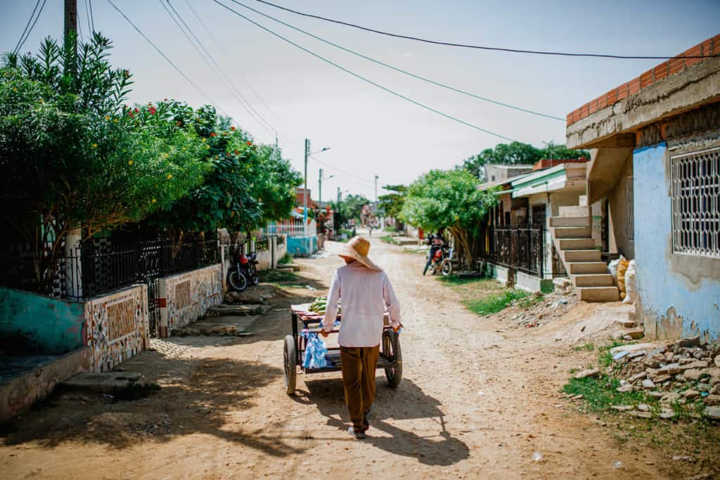 A person in a white shirt and a straw hat is pushing a cart down a dirt and gravel road.