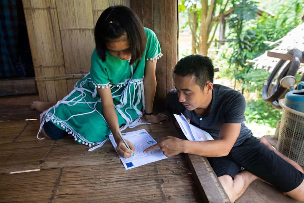 A Compassion staff member in Thailand helps a girl wearing a green and white dress to write a letter.
