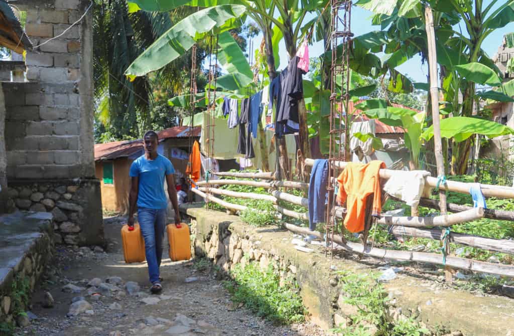 Young man wearing a blue shirt and jeans. He is carrying two large yellow containers full of water home for his family. There are clothes hanging on a clothesline next to him.