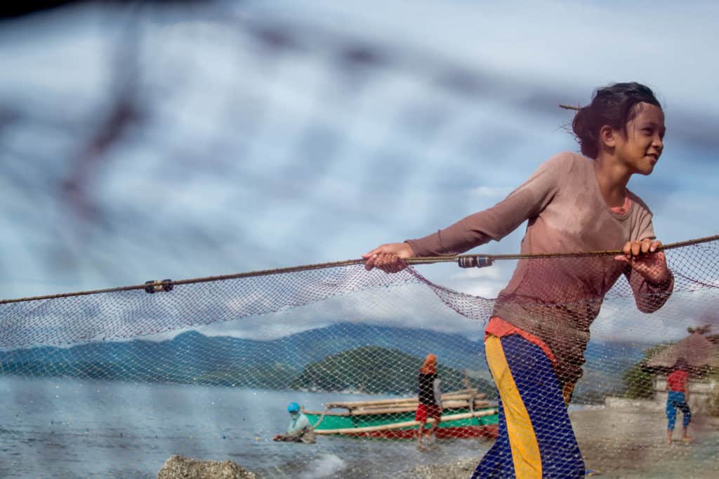 Ariane is wearing a brown shirt and purple pants. She is pulling a fishing net in to help her parents.