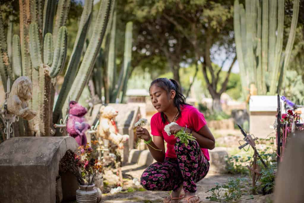 Melani is wearing a bright pink shirt with black floral print pants. She is kneeling down beside a headstone in the cemetary where she works. She is holding flowers.