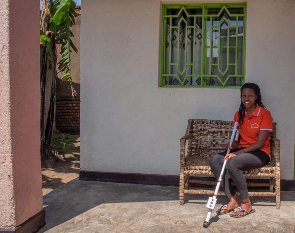 A young woman sits on a wicker chair holding the digital cane she invented. she is smiling and wearing an orange shirt.