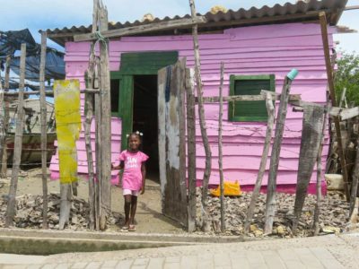 A girl wearing pink stands outside a small pink home with a tin roof. There is a fence made of pieces of wood and barbed wire around the home.