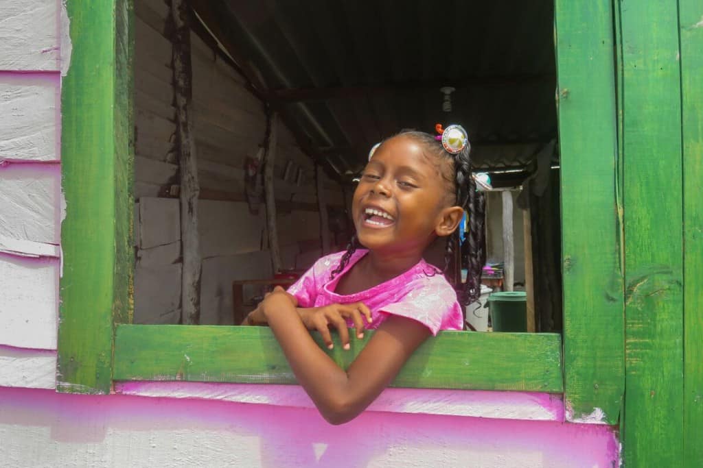 A young girl leans out the window of a pink house with a green windowsill. She is smiling and wearing a pink shirt