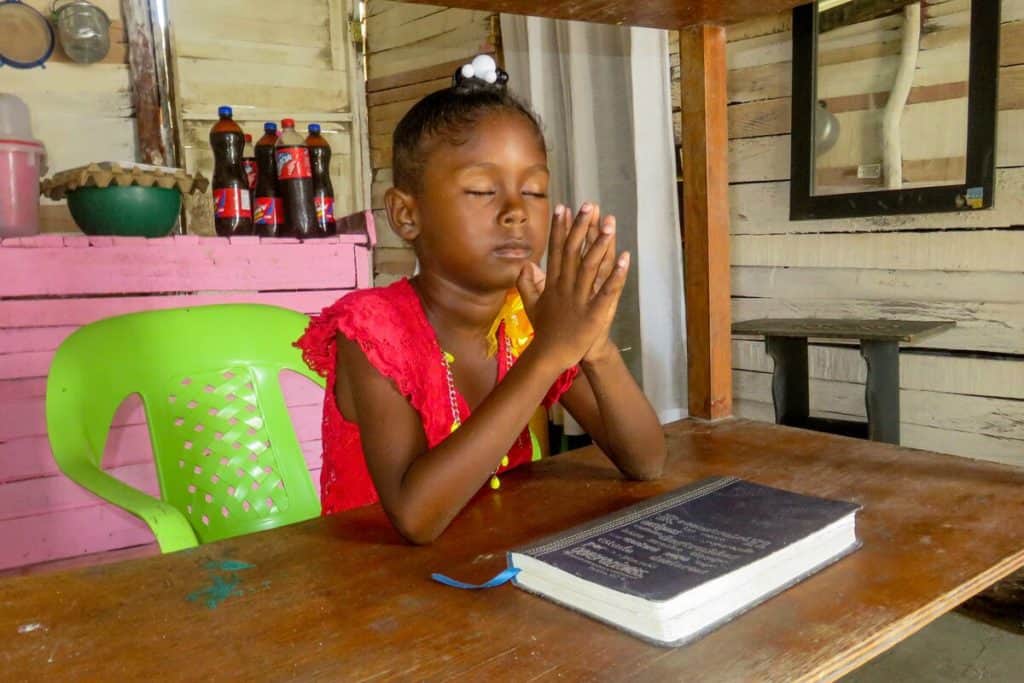 A girl sitting in a green plastic chair closes her eyes and presses her hands together in prayer. She is sitting in her new home at a table, where a Bible sits in front of her. 