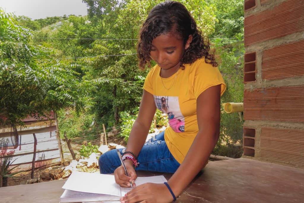 Rosa is wearing jeans and a yellow shirt. She is sitting outside her home and is doing the activities in her education guide from school.