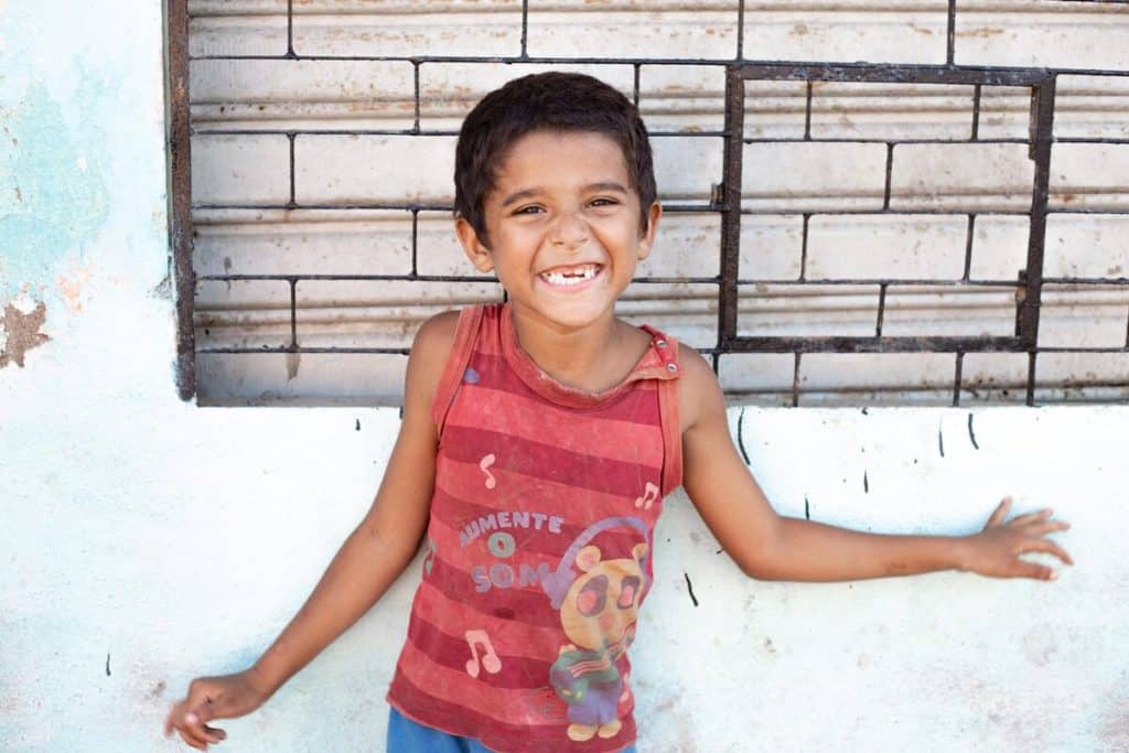 A Brazilian child wearing a red shirt smiles in front of a wall.