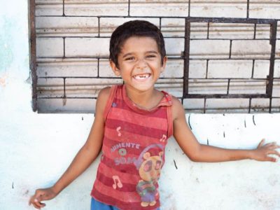 A Brazilian child wearing a red shirt smiles and stands in front of a wall