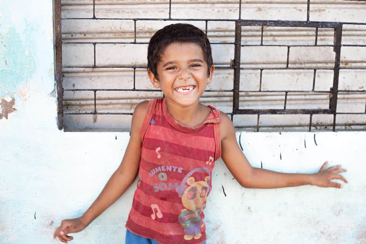 A Brazilian child wearing a red shirt smiles and stands in front of a wall