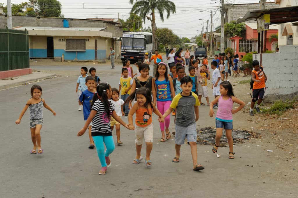 Young boys and girls walk smiling and laughing, some holding hands together, as they stroll as a large group on a paved street in their neighborhood in Ecuador. A bus is no the road behind them near various buildings on either side of the street.