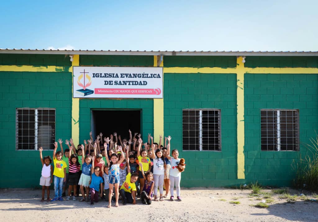 Classmates standing in front of the church. It is a green building with yellow accents. The children are all raising their hands.