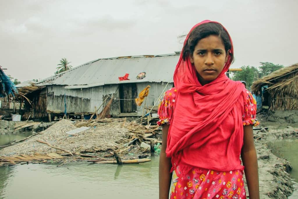 A pile of leaflike rubble behind Aduri, in front of the home made from tin sheets is all that remains from Aduri’s home. Aduri is wearing red.