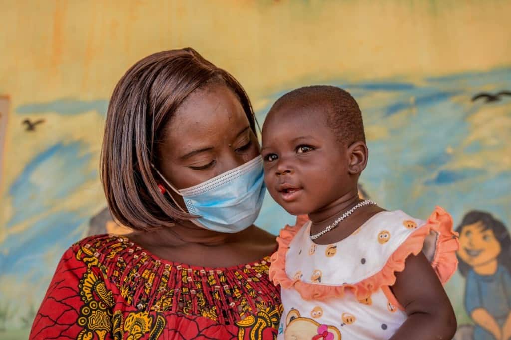 Farida is wearing an orange and white dress. Beatrice, wearing a red and yellow dress, is holding Farida in front of a mural inside the Compassion center.