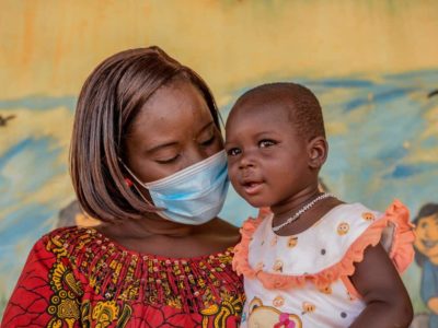 Farida is wearing an orange and white dress. Beatrice, wearing a red and yellow dress, is holding Farida in front of a mural inside the Compassion center.