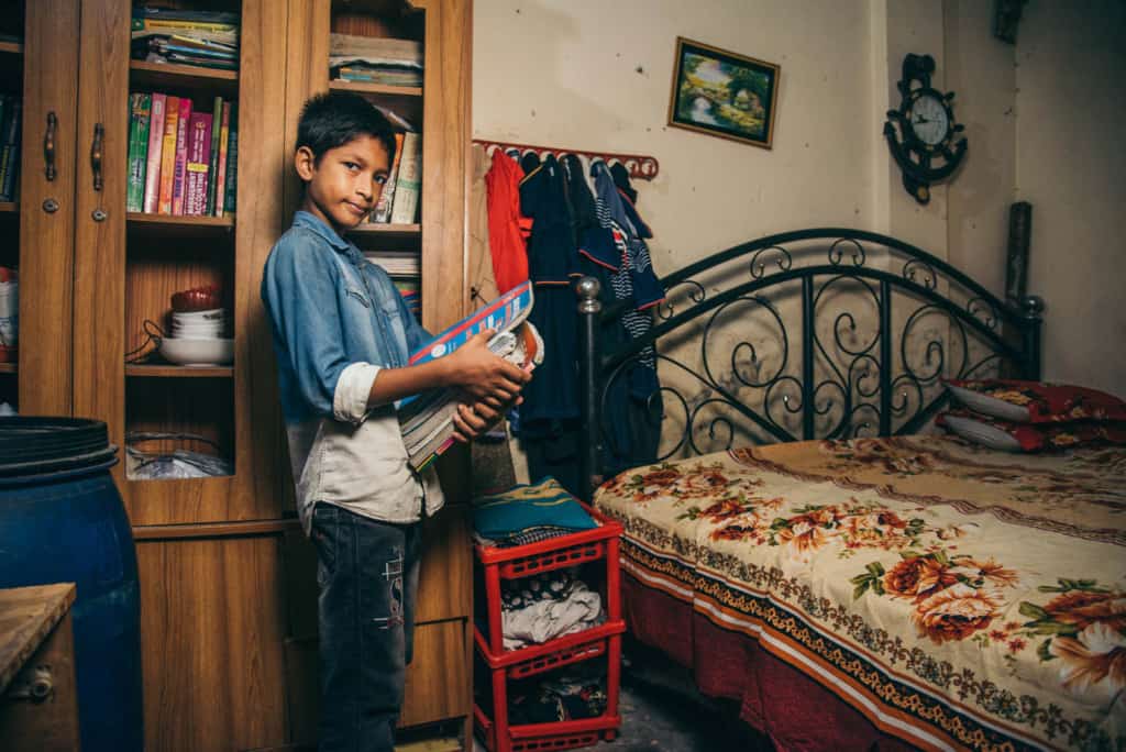 Boy wearing a denim shirt and jeans. He is standing in his room and behind him is a large cabinet full of his older siblings' books. He is holding an armful of books and next to him is his bed.