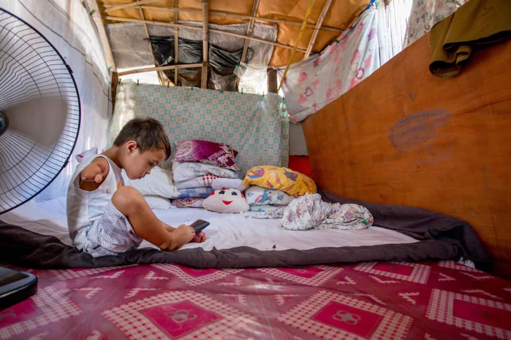 Jasper, wearing a white shirt and gray and white striped shorts, is sitting on his bed at home and he is using a cell phone with his feet. In the foreground of the picture is a fan. Behind his is a blue and white sheet hanging up as a room separator.