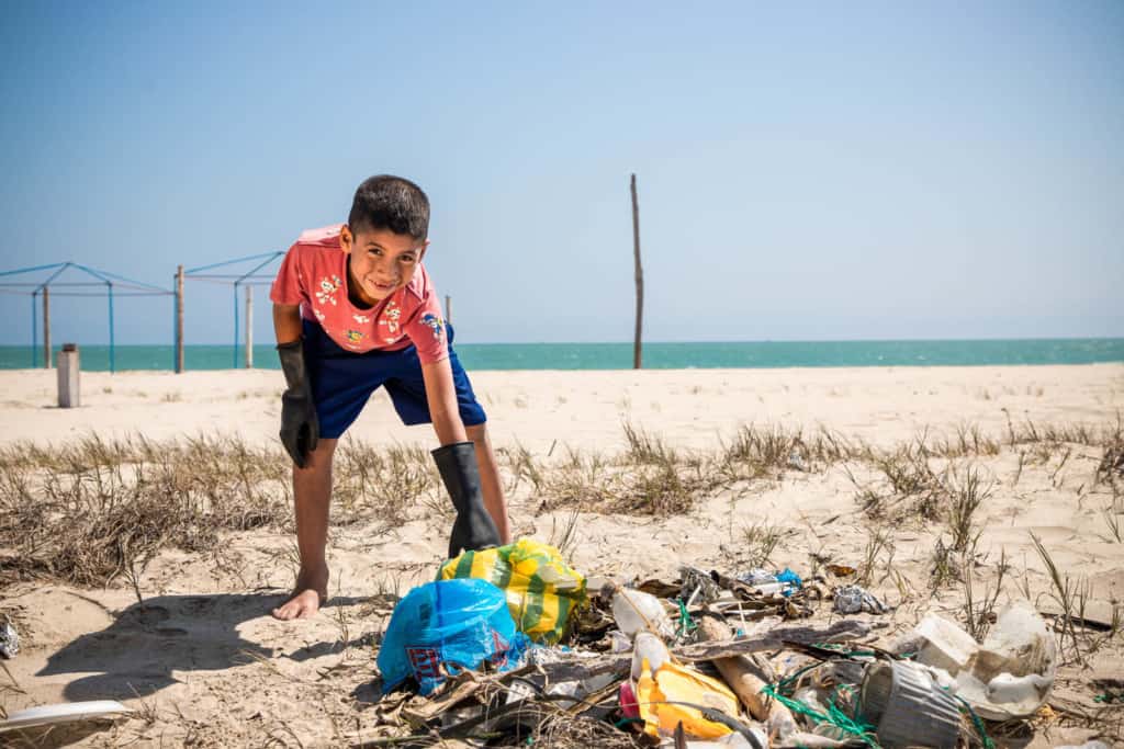 Boy wearing black shorts and a red shirt. He is leaning over and picking up trash.